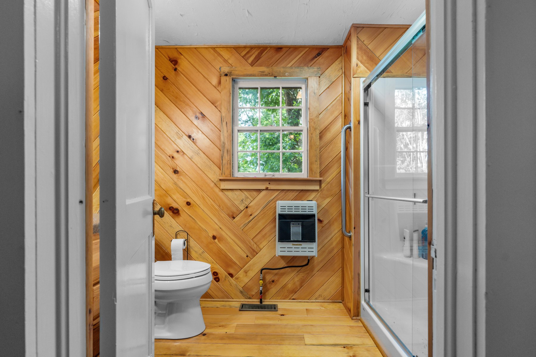 Cottage bathroom with wood-lined walls, a glass-enclosed shower, and a window facing the trees
