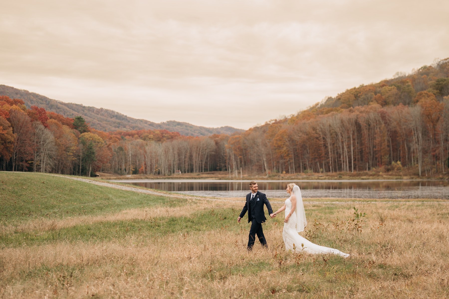 A bride and groom walking beside the lake with autumn forest behind them